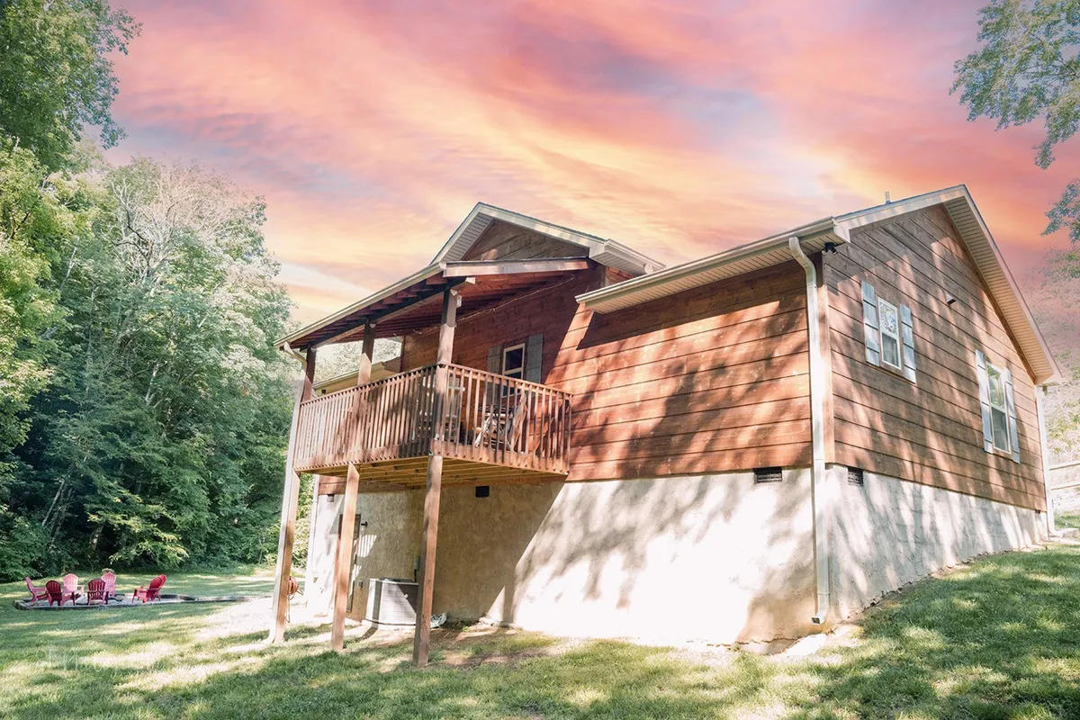 An inviting view from the wooden balcony, framed by lush woods and a colorful sunset sky. The outdoor seating area and glimpses of a fire pit create an atmosphere ideal for relaxing evenings and storytelling with loved ones.