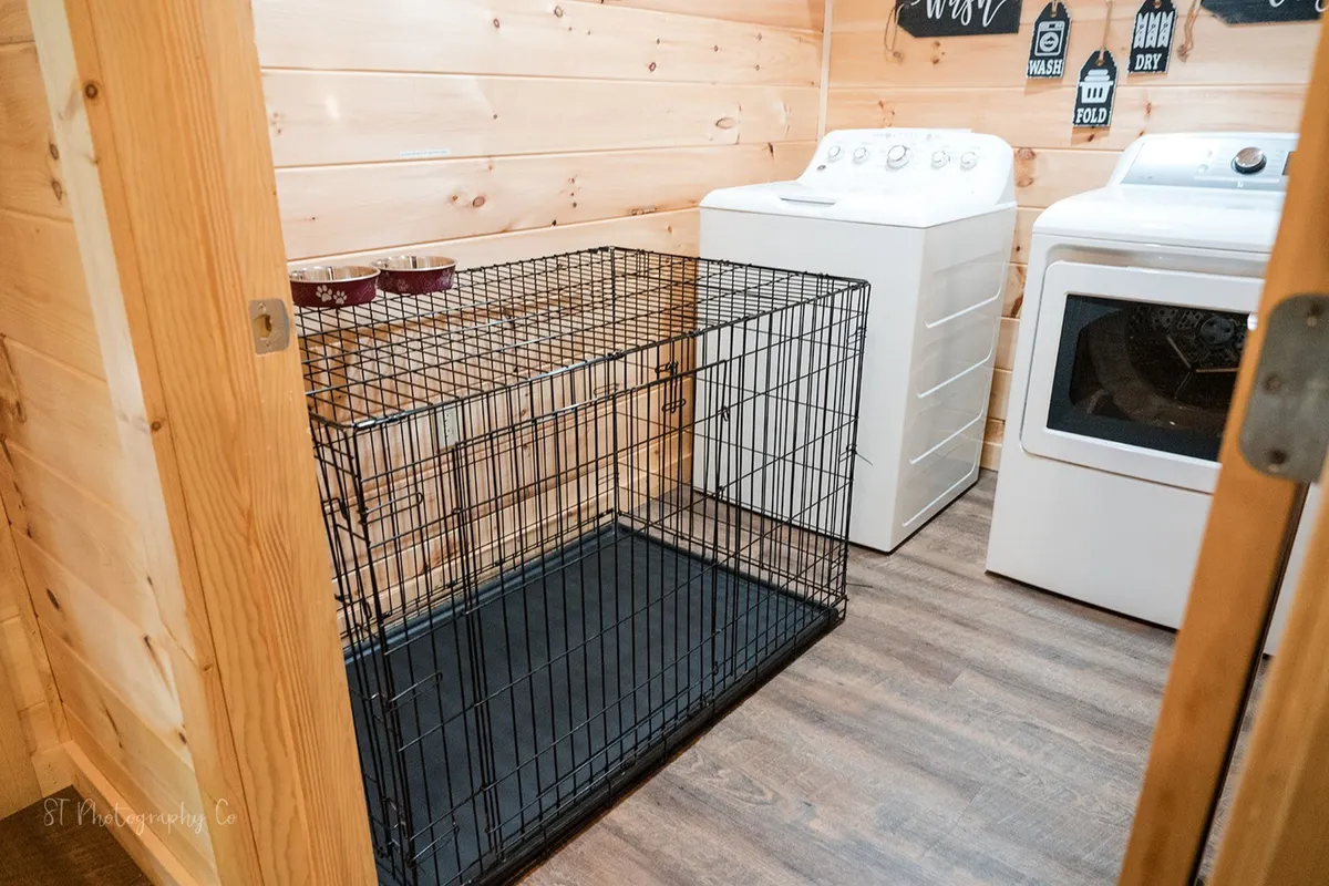 A practical interior image featuring a washing machine, dryer, and a large dog crate tucked against wood-paneled walls. The space is functional and homey, showcasing the thoughtful pet amenities provided by the cabin.