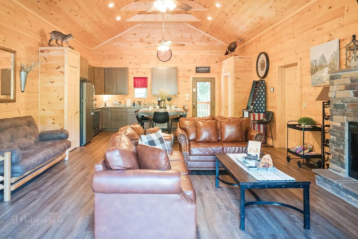 A cozy living room scene featuring leather sofas facing a striking stone fireplace, a large wooden dining table nearby, and sunlight streaming in from expansive windows. The space is open and ideal for group gatherings or quiet relaxation.