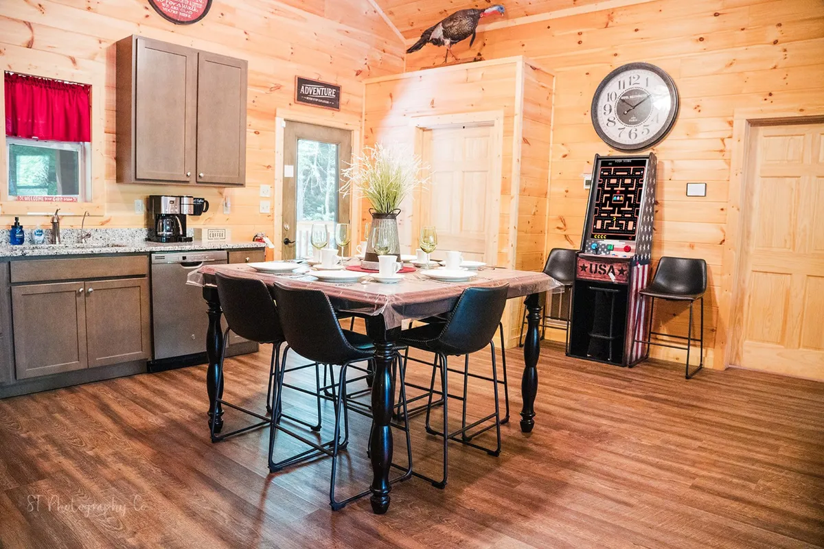 A cozy, rustic dining area inside the cabin features a large table, wooden kitchen cabinetry, a decorative wall clock, and a potted plant. The space is set up for family meals and togetherness after a day of adventure.