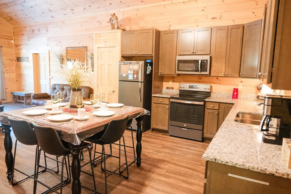A cozy interior view of a dining table set with plates and surrounded by modern kitchen appliances and warm wooden cabinetry. The scene suggests a welcoming environment for sharing meals and cooking together.