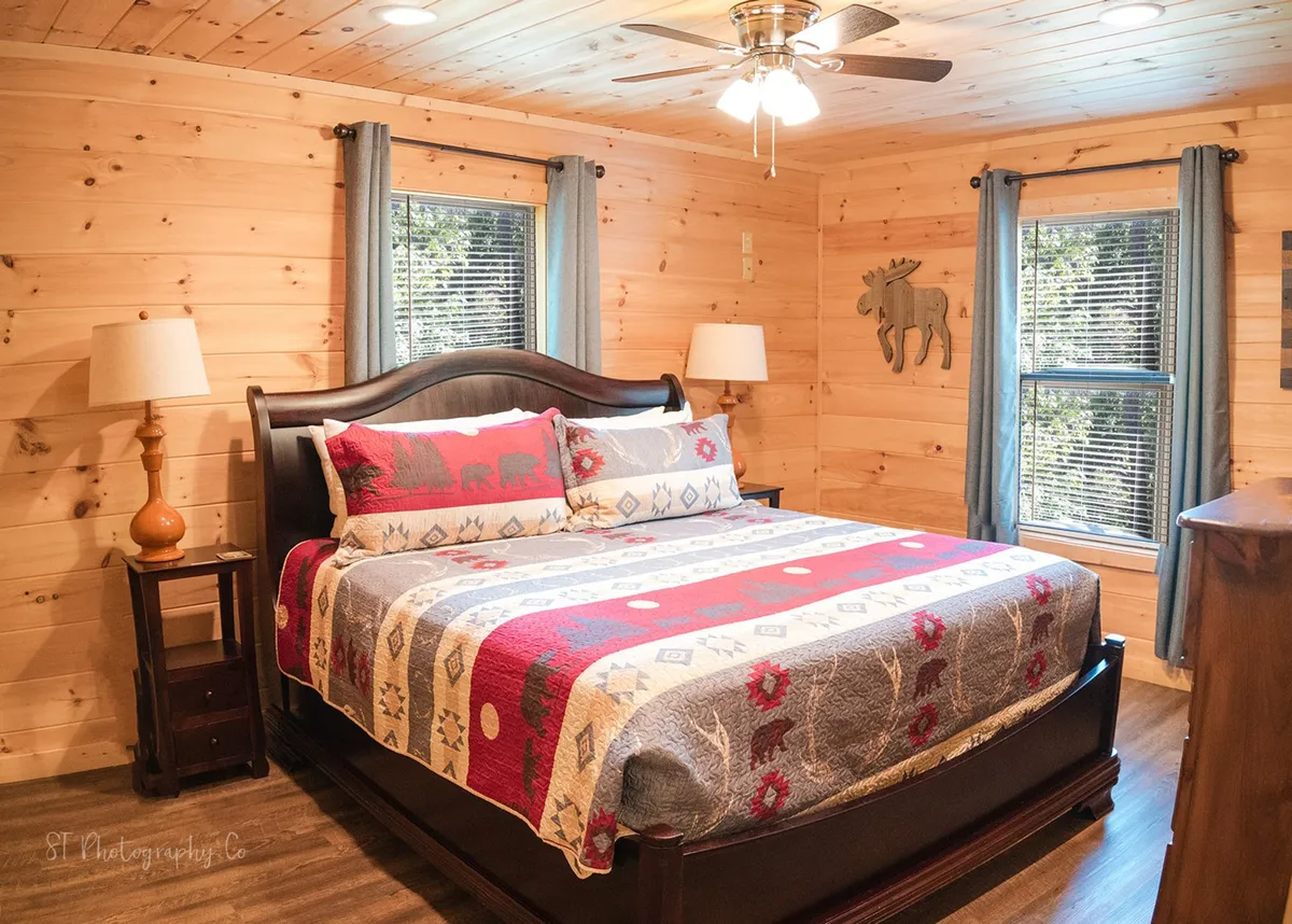 An inviting cabin bedroom with a warm, rustic vibe, featuring a bed with patterned bedding, wooden furniture, and windows with curtains that let in soft natural light. Moose wall decor adds a playful mountain touch.