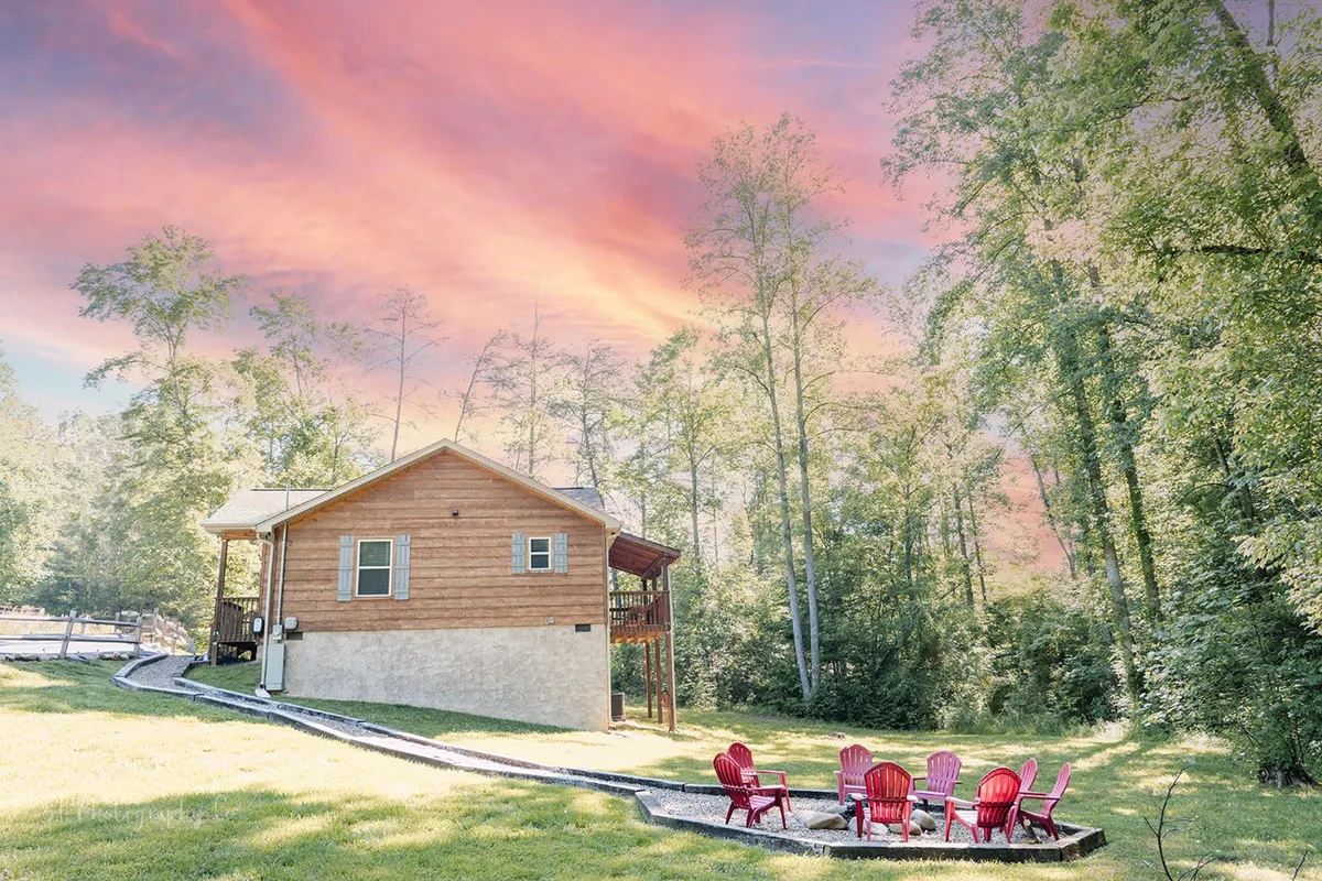 This exterior image displays the charming wooden cabin surrounded by lush greenery. Red Adirondack chairs and a pathway lead to a cozy fire pit, while the outdoor area invites families to relax, share meals, or gather around the fire.