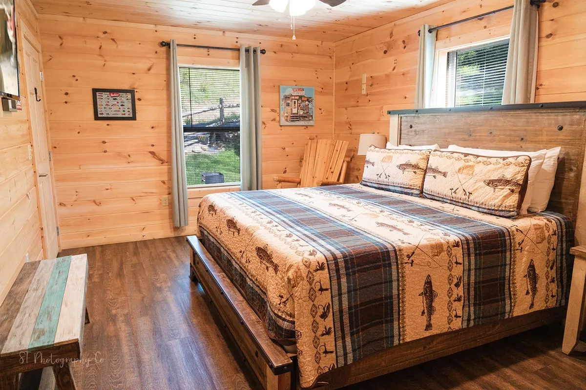 A cozy bedroom featuring a bed with a patterned quilt, rustic wooden furniture, and a large window offering a serene view of the outdoors. Sunlight streams in, inviting guests to relax and connect with nature from the comfort of the cabin.