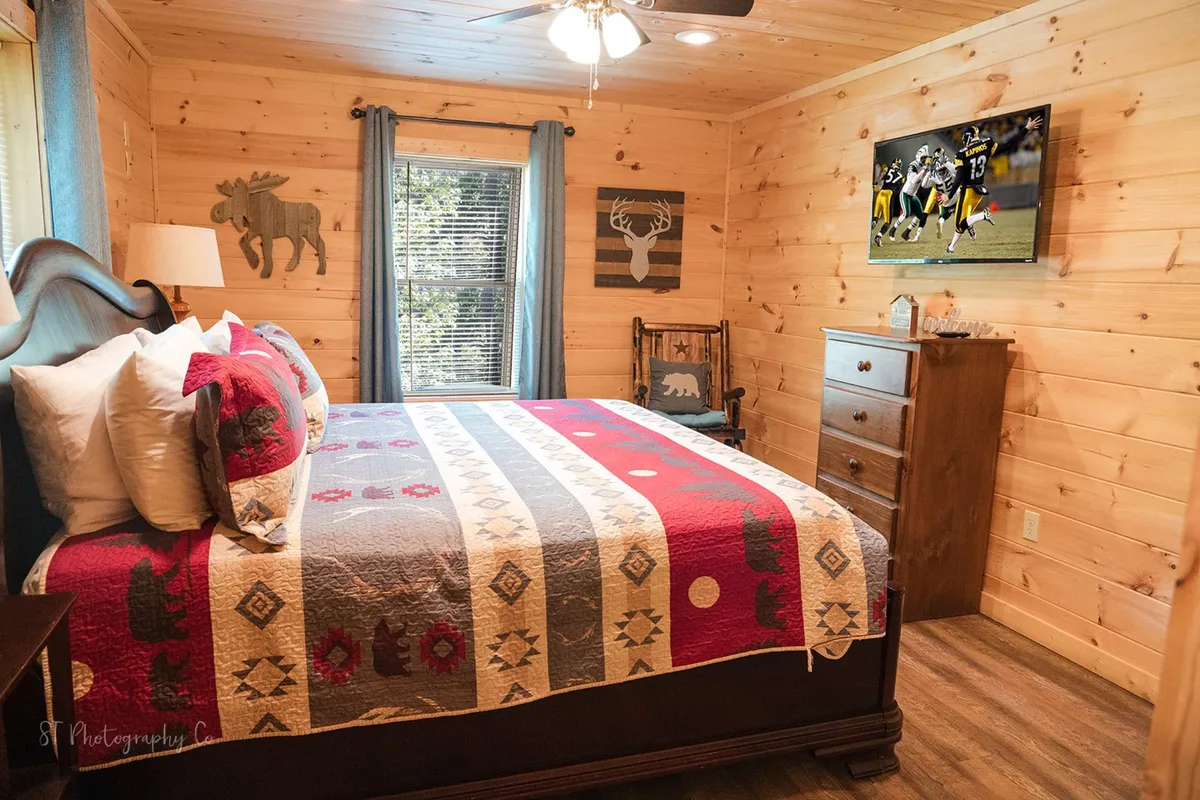 A king bedroom with a bed adorned with a patterned quilt, rustic wooden furniture, and a wall-mounted TV. The walls display decorative moose and deer art, contributing to the cabin's rustic charm. The room feels both comfortable and authentic to the mountain setting.