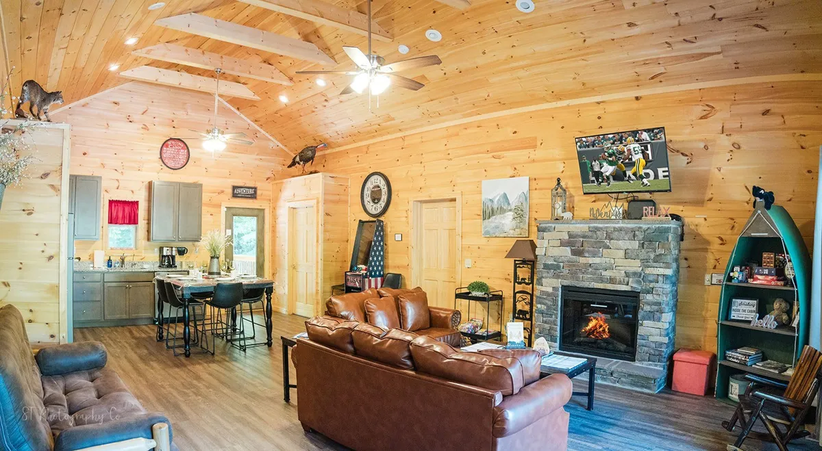 This image captures an open-concept living area with a plush sofa facing a modern fireplace, a wall-mounted television, and an adjoining dining area with a view into the kitchen. The space is brightened by natural sunlight pouring in through large windows, and the arrangement invites gatherings and relaxation.