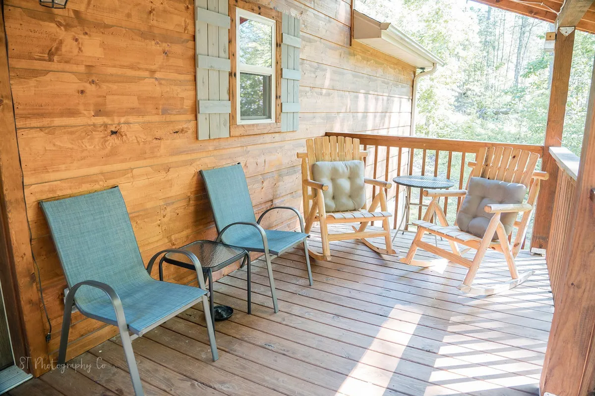This exterior image displays a charming wooden deck with rocking chairs and a small table, framed by lush greenery. The cozy seating area invites relaxation and offers a view of the surrounding natural landscape, perfectly suited for unwinding outdoors with a pet by your side.
