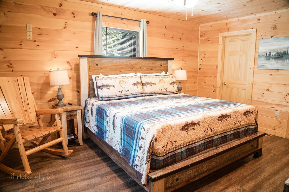 The image features a cozy bedroom with a patterned quilt on the bed, a wooden rocking chair beside a table lamp, and framed landscape artwork on the wood-paneled wall. Sunlight filters through a nearby window, enhancing the feeling of warmth and tranquility in the space.