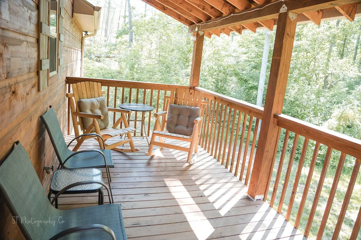 A covered porch with wooden railing, featuring a pair of rocking chairs and a small table. The setting overlooks lush greenery, inviting guests to relax and take in the morning serenity or enjoy peaceful evenings.