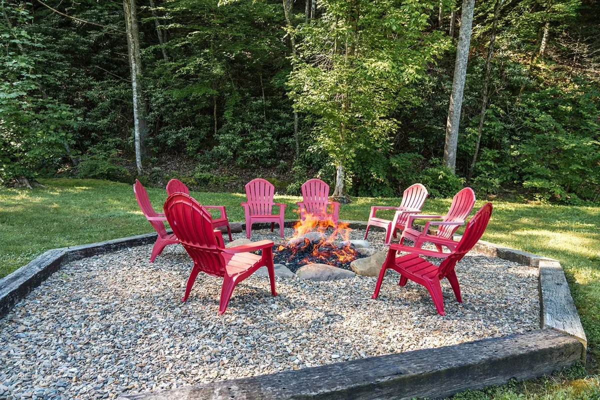This exterior image features a fire pit encircled by vibrant red chairs, set amidst lush greenery. The scene is outdoors, with easy access implied by the open and level area leading up to the seating arrangement.