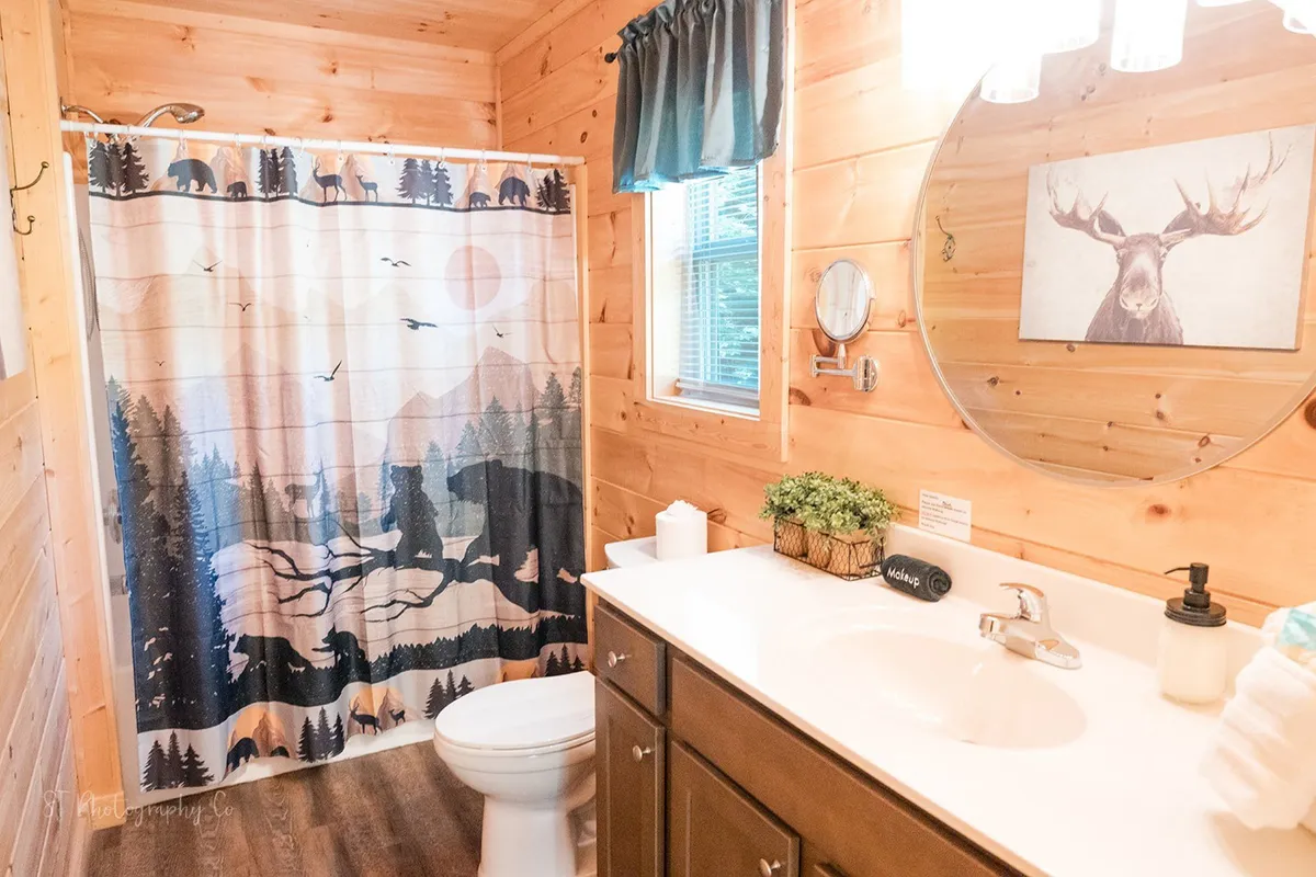 A bathroom with a shower curtain, vanity area, framed wall art, and a window allowing natural light to flow in. The decor is warm and inviting, with nature-inspired accents that echo the cabin’s overall theme.